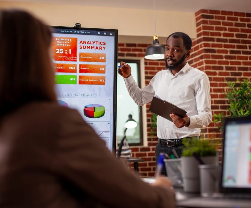Businessman with clipboard in hand, using digital monitor to explain analytics summary in meeting. African american entrepreneur doing project presentation, planning marketing strategy with his team.